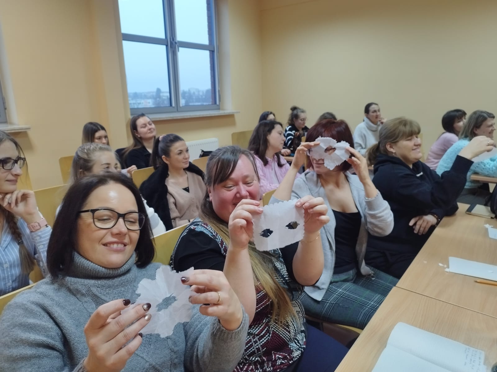 Studentki Pedagogiki prezentują wycinane z papieru prace. W tle uśmiechnięte studentki.